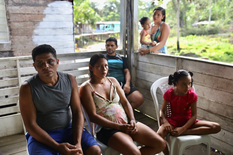 Los residentes se sientan en una terraza de madera y algunos cargan a niños pequeños.  La vegetación de la selva amazónica es visible más allá de la cubierta.