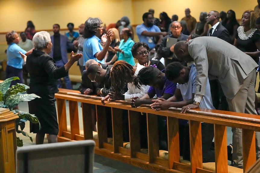 Personas arrodilladas detrás de un banco con la cabeza inclinada orando en la iglesia y otras personas reunidas a su alrededor