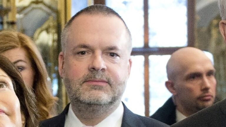 A man with grey stubble looks at the camera as he stands among a group of people.