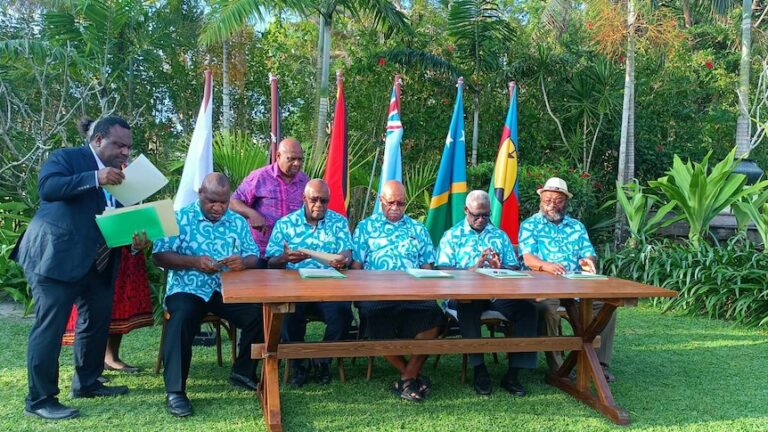 Pacific leaders wearing matching blue Hawaiian shirts sit at table with flags in the background.