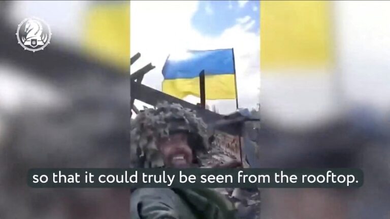 A soldier wearing a camouflage taking a selfie from a low angle with the blue and yellow ukraine flag behind him on a rooftop