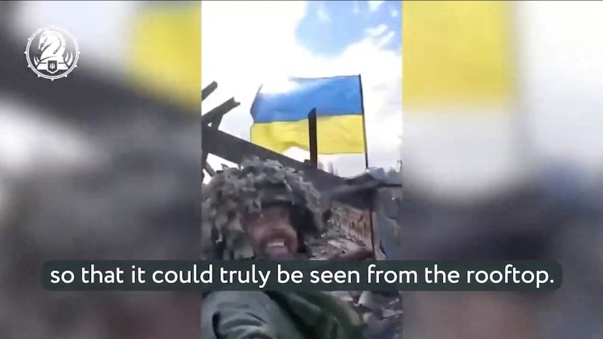 A soldier wearing a camouflage taking a selfie from a low angle with the blue and yellow ukraine flag behind him on a rooftop