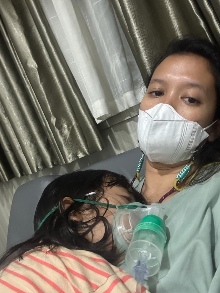 Una niña pequeña, con una máscara de oxígeno sobre la nariz y la boca, duerme sobre el pecho de su madre.