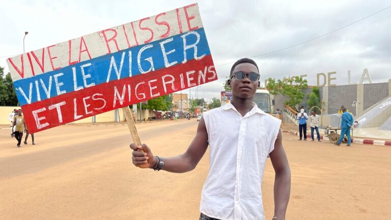 Man in white shirt and sunglasses looks serious while holding sign that says "Long Live Russia, Long Live Niger and Nigeriens".