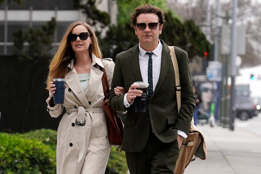 Un hombre y una mujer con ropa corporativa y gafas de sol de gran tamaño caminando por una calle sosteniendo un café. 