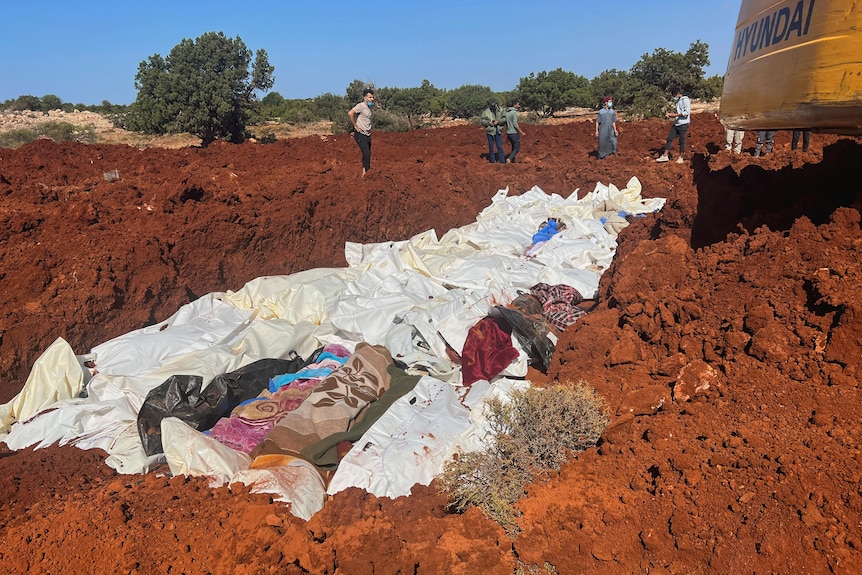 Los cuerpos envueltos en mantas son colocados en una fosa común; en la esquina superior derecha se puede ver una excavadora.