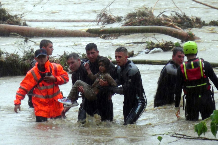 Hombres con trajes de neopreno cargan a una niña a través de aguas inundadas que les llegan hasta los muslos. 