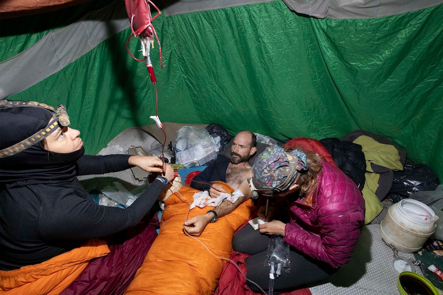 Un hombre acostado en un saco de dormir con un suero intravenoso dentro de una tienda de campaña con dos expertos médicos en equipo para actividades al aire libre y faros.
