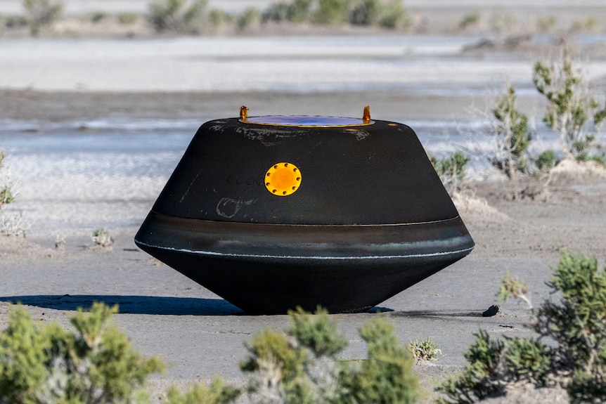 Una cápsula de color gris oscuro con una puerta amarilla se encuentra en el suelo del desierto.