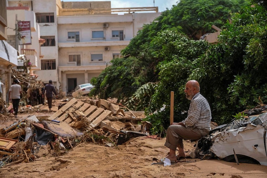 Un anciano sentado en un automóvil dañado, rodeado de escombros de la inundación, con edificios de varios pisos al fondo.