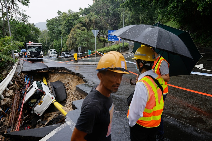 Trabajadores con cascos amarillos y sombrillas negras que rodean un gran agujero en una carretera con un automóvil blanco dentro