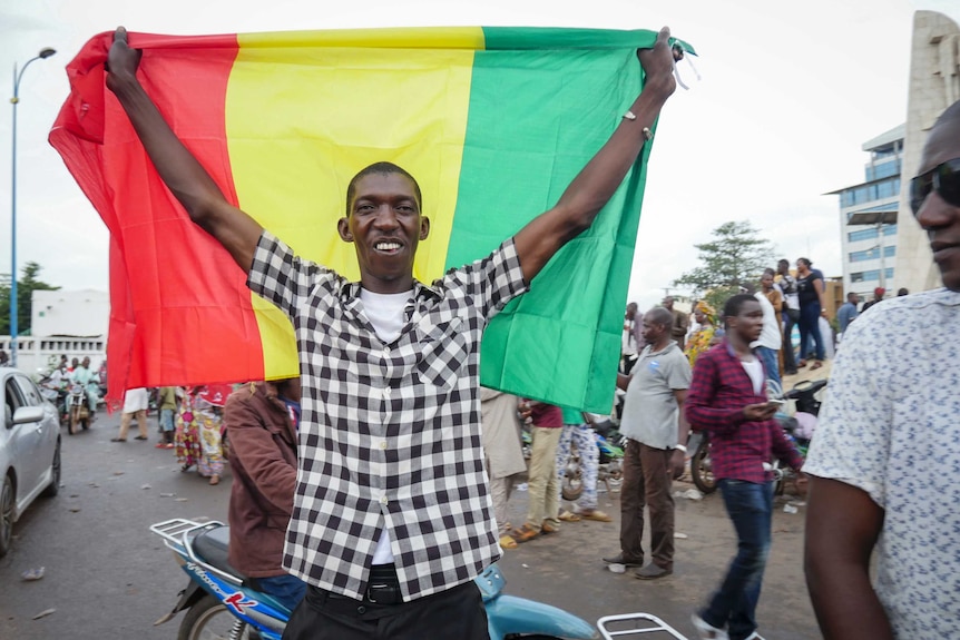 Un hombre negro delgado y sonriente sostiene una bandera roja, amarilla y verde en una concurrida calle africana.