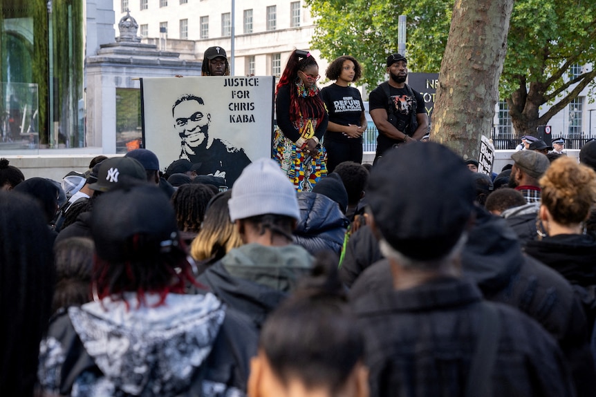 Cuatro manifestantes negros suben al escenario frente a una multitud, uno de ellos sostiene un cartel que dice "justicia para Chris Kaba"