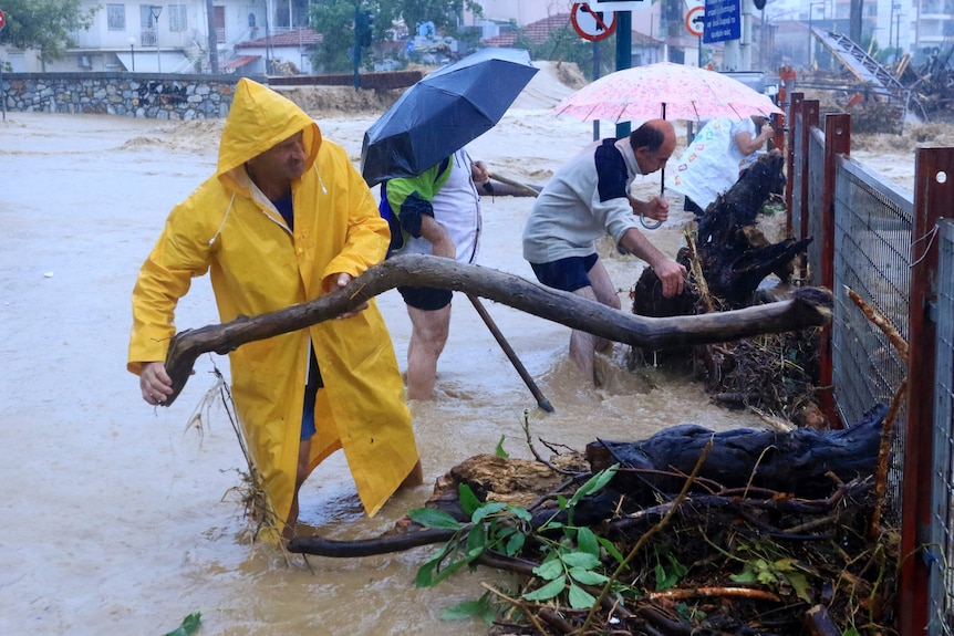 Un hombre con un impermeable amarillo levantando una rama del agua hasta la altura de las pantorrillas. 