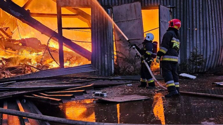 Two firefighter stand holding a house and spraying water onto a building that is engulfed in bright orange flames.