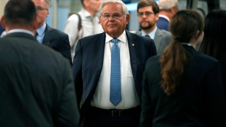 An US senator wearing black suit and blue tie surrounded by people.