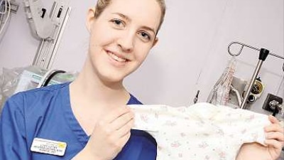 A young woman wearing a blue nurse's uniform smiles while holding up a babygrow