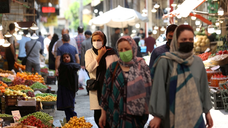A group of women wearing hijabs and masks walk through a colourful outdoor market.