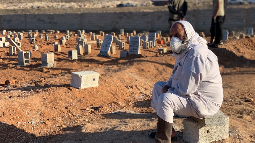 man wearing white personal protective equipment sits while numerous makeshift gravestones can be seen behind him