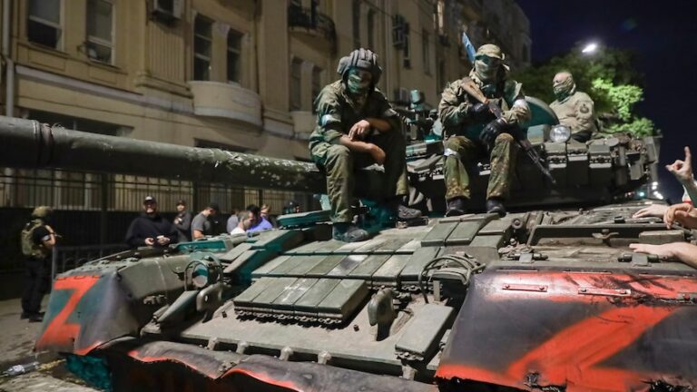 Three men in military uniforms and face masks sit on a tank on Russian street at night. The tank bears the letter Z in red.