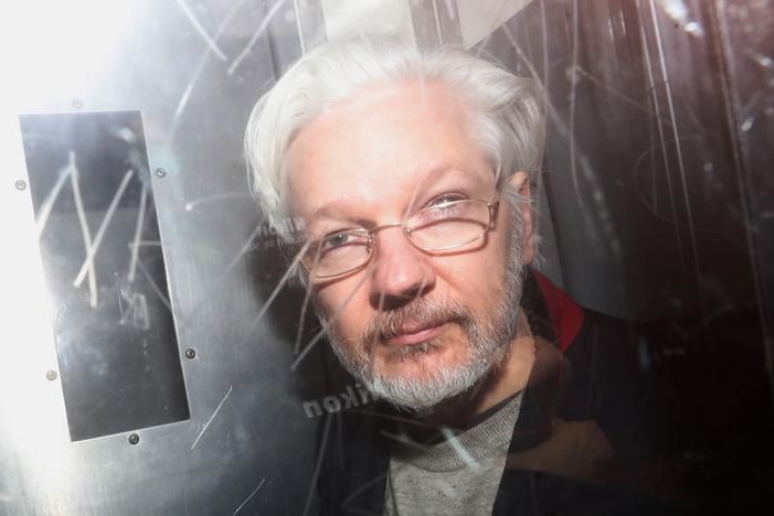 Julian Assange mira a la cámara mientras lo fotografían detrás de un vidrio con graffitis grabados.  Sus canas han vuelto.