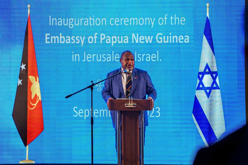 El hombre se encuentra en el podio entre las banderas de PNG e Israel frente a un fondo azul. 