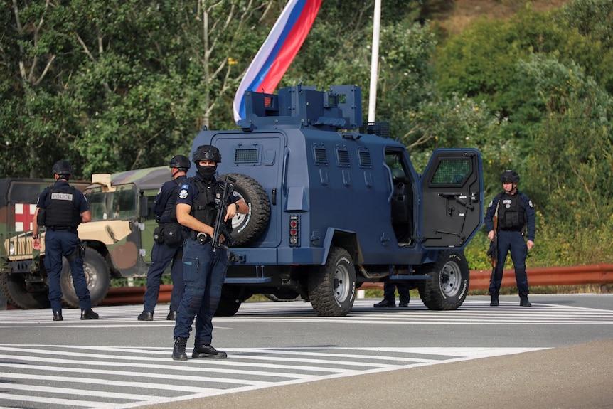 Agentes de policía con rifles y cubiertas para la cara se encuentran en un cruce cerca de un vehículo blindado azul.