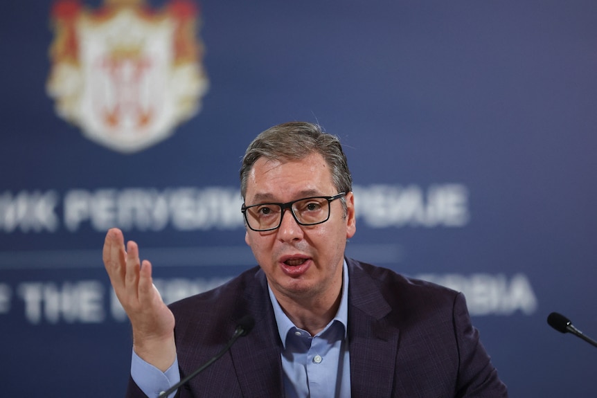 Un hombre blanco de mediana edad con gafas y traje hace gestos mientras habla por un micrófono frente a un fondo azul.