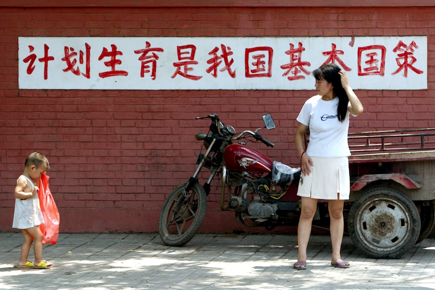 Una joven madre china observa a su hijo caminar frente a un cartel que dice "El control de la natalidad es una política estatal básica de nuestro país.".