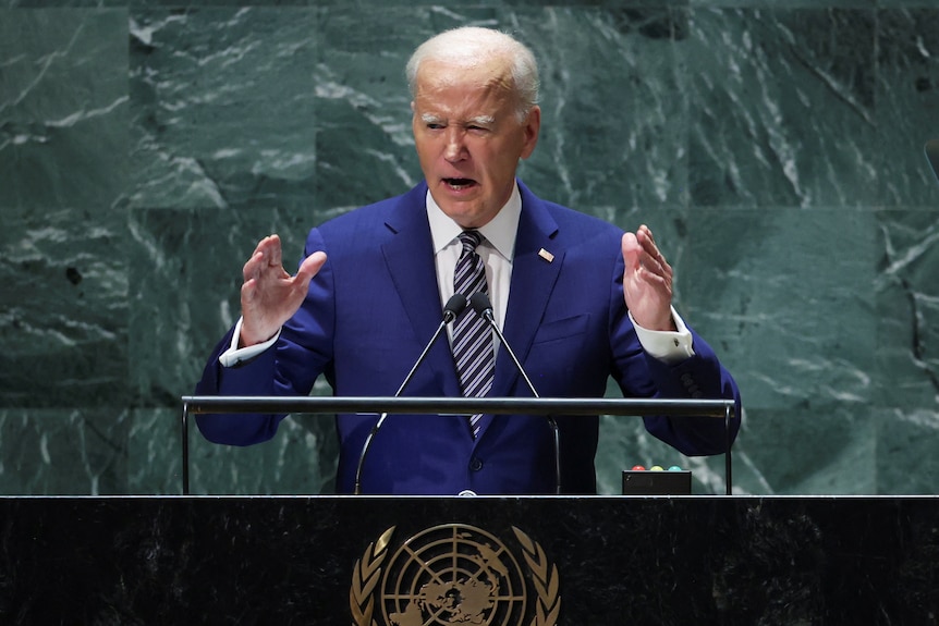 Joe Biden hace gestos con las manos mientras está de pie en el podio y habla.  Viste un traje azul y corbata a rayas.