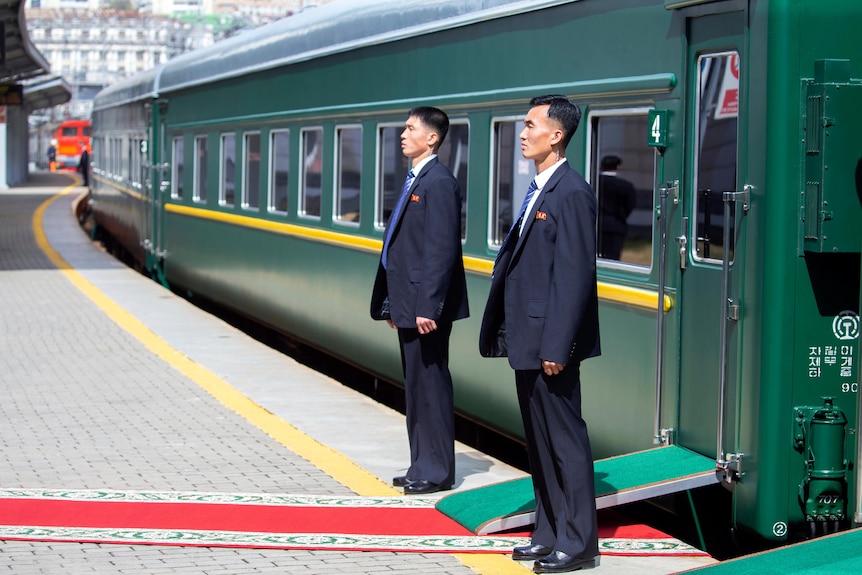Dos guardias de seguridad se encuentran en posición de firmes frente a la puerta de un tren verde en una estación de tren.