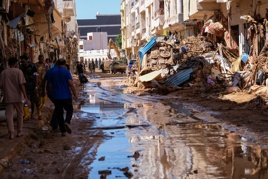 La gente camina en el barro entre edificios destruidos, una excavadora acumula escombros