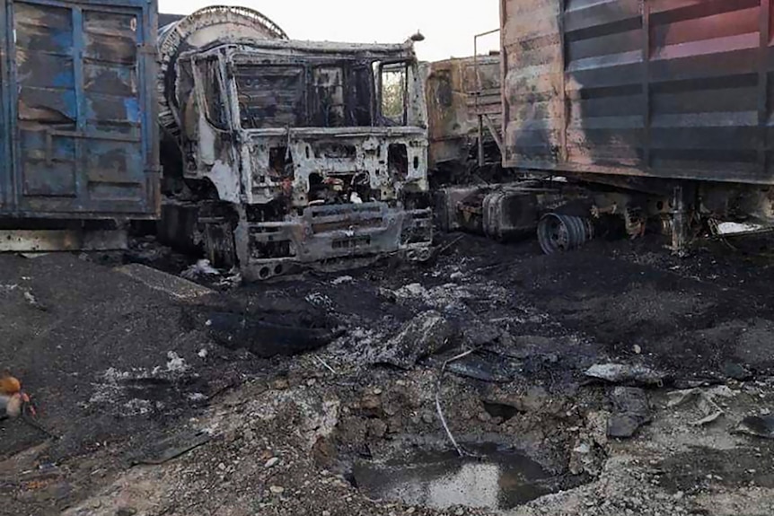 Una escena mediana de camiones destruidos y quemados, junto a un contenedor dañado y montones de tierra y escombros.