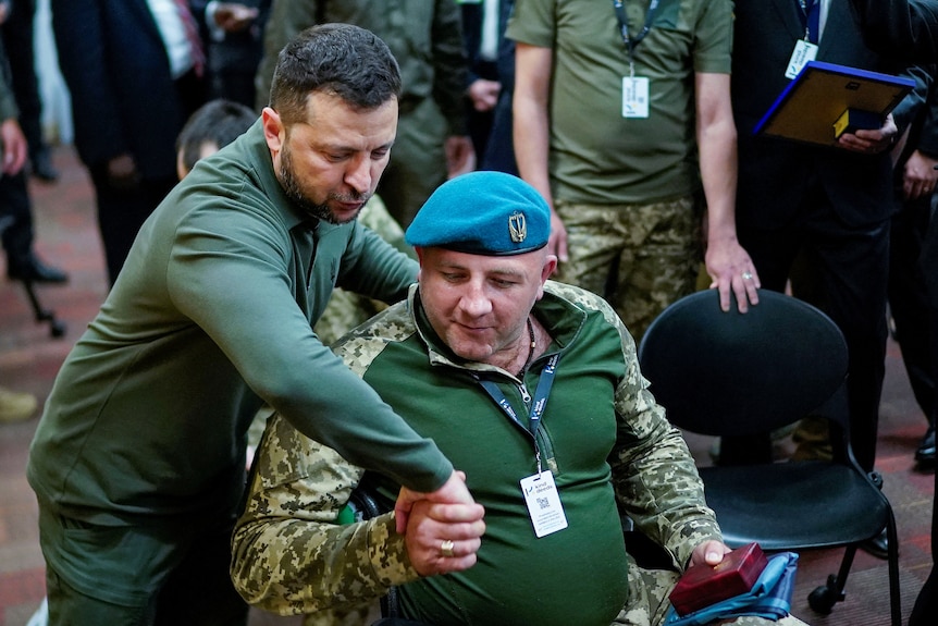Volodymyr Zelenskyy le da la mano a un soldado herido que está sentado.