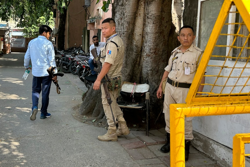 Tres hombres armados, dos de ellos uniformados, se encuentran junto a un gran árbol junto a un edificio, detrás de una valla metálica.