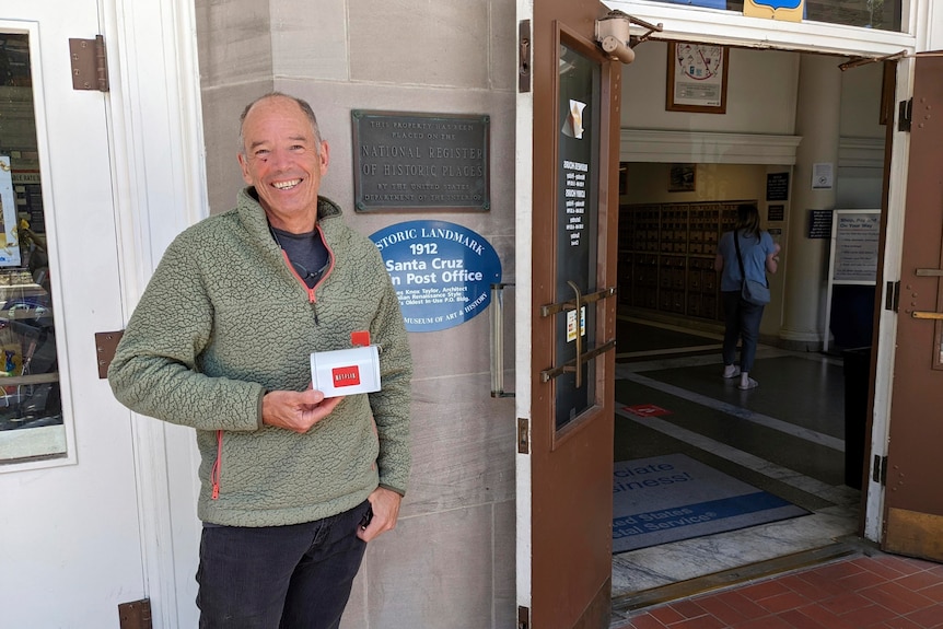 Un hombre sonriente sostiene un buzón con la marca Netflix frente a la entrada de una oficina de correos.