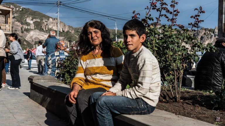 A woman and her son sit on a bench near a tree in the sun.