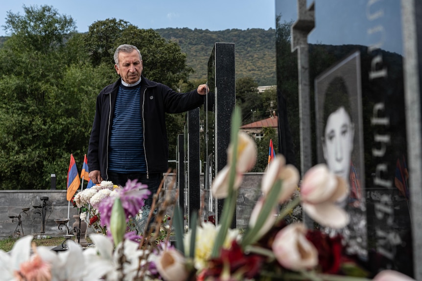 Un hombre vestido con ropa oscura mira una hilera de tumbas bordeadas de flores.