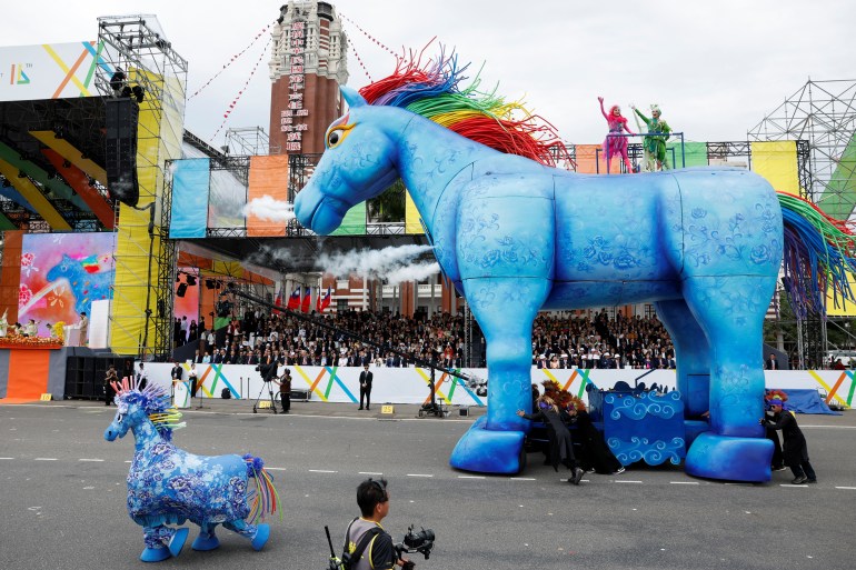 Una efigie gigante de un caballo azul con una melena arcoíris participando en la ceremonia de inauguración.  Un caballo azul más pequeño está al lado y hay una multitud detrás.