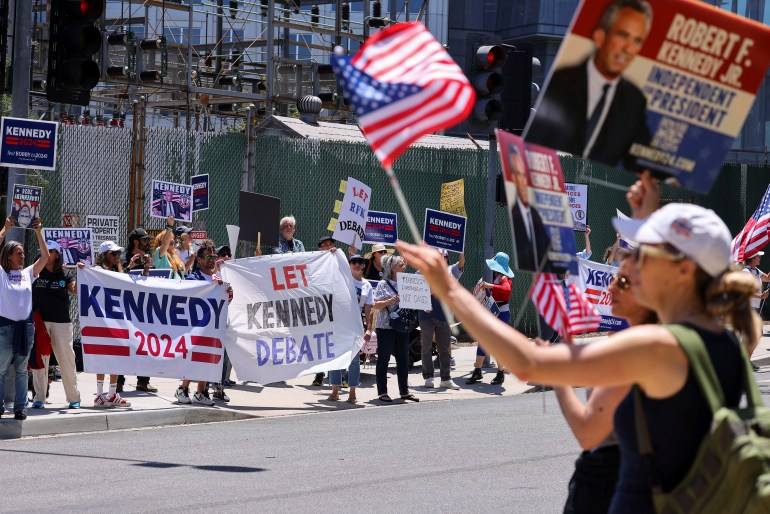 Una mujer ondea una bandera estadounidense en un mitin de Robert F. Kennedy Jr, donde otra persona sostiene una pancarta que dice: "Dejemos que Kennedy debata"