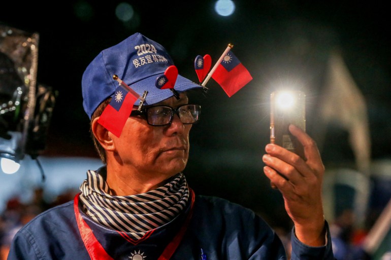 Un partidario del KMT en un mitin.  Es un hombre mayor y lleva una gorra azul decorada con pequeñas banderas y corazones de Taiwán. 