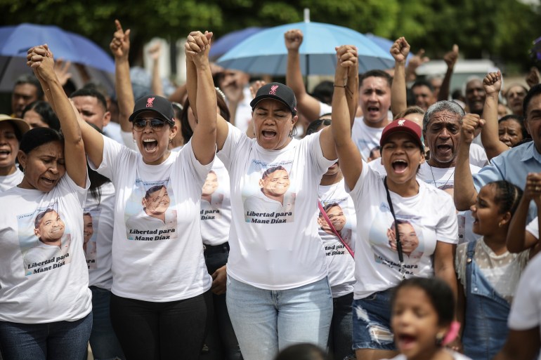 Mujeres con gorras y camisetas se cruzan y levantan los brazos en el aire mientras protestan para presionar por la liberación del padre de Luis Díaz.  La cara del padre está impresa en cada una de las camisetas.