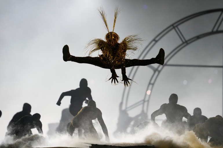 Los bailarines actúan en la ceremonia de clausura en París. Uno está dando un salto de altura en el aire. Otros están agachados. Hay mucho humo blanco.